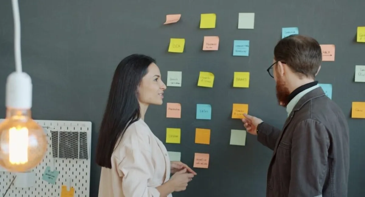 Two colleagues discussing sticky notes on an office wall, the planning work behind the prompt sets that AI tools for brand visibility 2025 2026 track