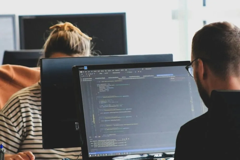 Two people working on code at monitors in a bright office, the debugging work that real-time analytics tools for AI inference are meant to support
